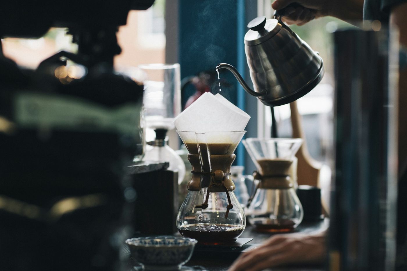 A barista pours from a kettle into a pour-over brewer above a glass carafe