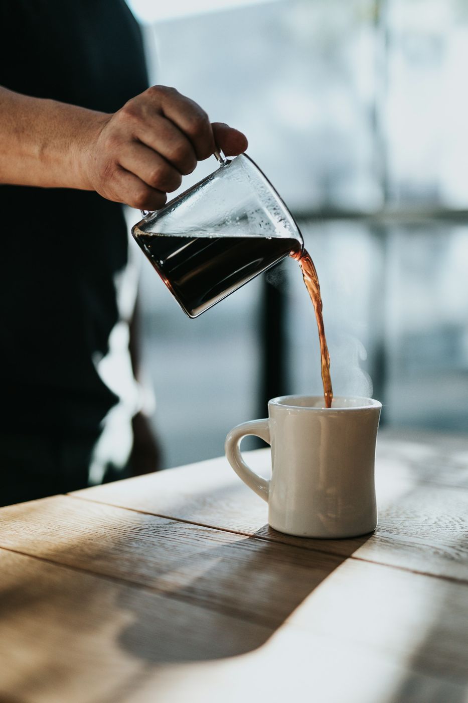 Black coffee poured from a glass server into a white mug on a wooden table